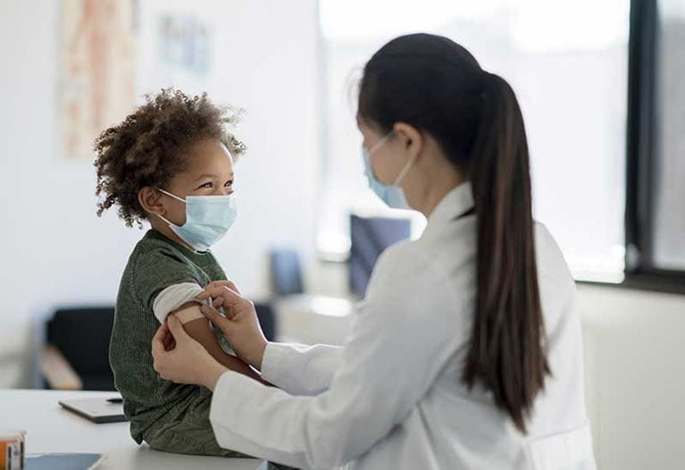 Boy wearing mask gets bandage placed on arm by female doctor. 
