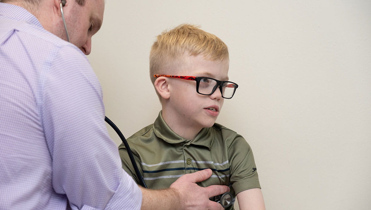 Smiling boy with blonde hair wearing a green shirt and eyeglasses.